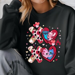 K&ouml;nnte beinhalten: Schwarzes Sweatshirt mit einem Valentinstagsdesign. Das Design zeigt zwei Paare von Zeichentrickfiguren in herzf&ouml;rmigen Rahmen mit den Buchstaben "X" und "O". Die Buchstaben haben ein rotes und goldenes Muster. Herzen und Sterne sind um das Design verstreut.