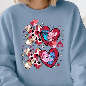 K&ouml;nnte beinhalten: Hellblaues Sweatshirt mit einem Valentinstagsdesign. Das Design zeigt die Buchstaben X und O, Herzen und die Zeichentrickfiguren Stitch und Angel. Die X und O haben ein rotes, goldenes und wei&szlig;es Muster.