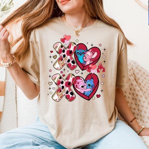 K&ouml;nnte beinhalten: Beiges T-Shirt mit einem Valentinstags-Design. Das Design umfasst zwei gro&szlig;e "XO"-Formen mit einem rosa und braunen Muster und zwei rote Herzformen mit Cartoon-Figuren. Zus&auml;tzliche Herzen, Sterne und ein Schloss sind ebenfalls enthalten.