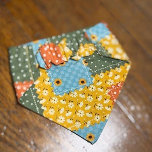 Puede incluir: Un pañuelo patchwork para mascotas con una mezcla de patrones florales y geométricos. El pañuelo presenta tonos amarillos, azules, verdes y naranjas, con un diseño de girasoles y margaritas. El pañuelo está doblado y listo para usar.