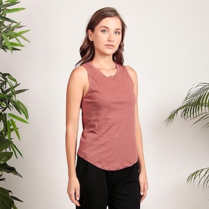 May include: A woman is wearing a rust-colored sleeveless tank top with a relaxed fit. The tank top has a round neckline and a slightly curved hemline. The woman is wearing black pants.