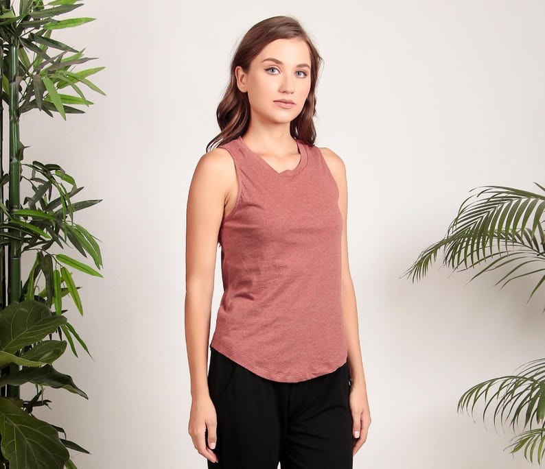 May include: A woman is wearing a rust-colored sleeveless tank top with a relaxed fit. The tank top has a round neckline and a slightly curved hemline. The woman is wearing black pants.
