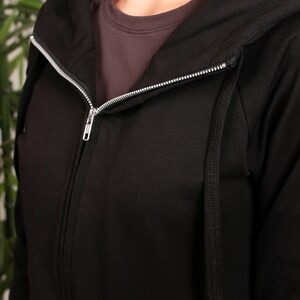 Puede incluir: Sudadera con capucha negra con cremallera y cord&oacute;n. La cremallera est&aacute; parcialmente abierta, revelando una camiseta interior gris.