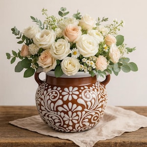 Puede incluir: Un arreglo floral de rosas blancas y melocotón, margaritas y vegetación en un jarrón de cerámica marrón. El jarrón tiene un patrón floral blanco y marrón y dos asas. El arreglo está sobre un paño de lino beige sobre una superficie de madera.