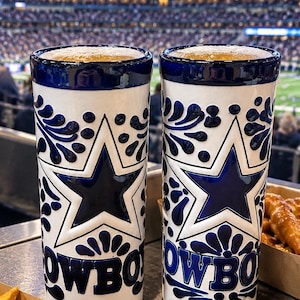 May include: Two tall ceramic tumblers featuring a white base with deep blue hand-painted floral patterns and a prominent blue star emblem. The text COWBOY is embossed in blue at the base of each cup. The rims are finished in a solid dark blue glaze.
