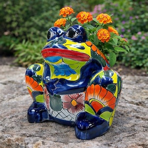 Könnte beinhalten: Ein handbemalter Keramik-Frosch-Pflanztopf mit leuchtenden Farben. Der Frosch hat einen blauen Körper, gelbe Akzente und orangefarbene und grüne Blumenmuster. Das Maul des Frosches ist rot und enthält eine Gruppe orangefarbener Blumen.
