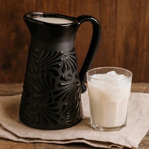 May include: A black ceramic jug with a handle and a floral pattern, filled with a white liquid. A clear glass with a white drink and ice sits beside the jug on a beige cloth.