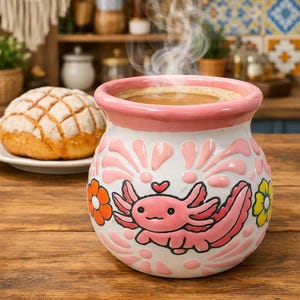 May include: A white and pink ceramic mug featuring a cartoon axolotl. The mug contains a hot drink, with steam rising from it. A concha pastry is on a plate. The mug has a pink rim and floral patterns.