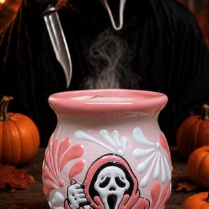 May include: A light pink ceramic cauldron mug featuring a white and pink embossed graphic of a masked figure holding a knife. The mug has a wide rim and is filled with a steaming liquid. The design includes stylized floral patterns in white and pink.