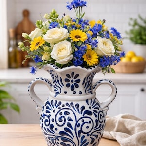 Puede incluir: Un jarrón de cerámica blanca con motivos florales azules contiene un vibrante ramo de rosas blancas, girasoles amarillos y acianos azules. El jarrón tiene dos asas y está sobre una mesa de madera, creando una exhibición brillante.