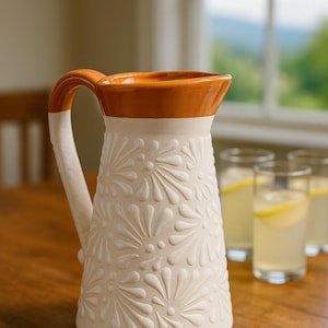 Puede incluir: Jarra de cerámica color crema con diseño floral en relieve. El borde y el asa son de color marrón anaranjado. La jarra está sobre una mesa de madera, con vasos de limonada al fondo. Un artículo decorativo y funcional.