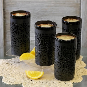 Op de afbeelding: Vier zwarte keramische bekers met een reliëf bloemmotief. Elke beker bevat een lichtgekleurde drank en een citroenschijfje. De bekers staan op een doorzichtig dienblad en een kanten kleedje. De bekers zijn ongeveer 15 cm hoog.