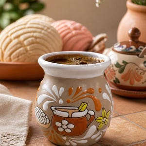 May include: A ceramic mug filled with coffee, featuring a brown and white design with orange and yellow accents. The mug is decorated with a stylized image of a pot and flowers. Two conchas and a decorative jar are in the background.