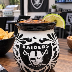 May include: A ceramic mug featuring a black and white Raiders logo with a shield design and decorative floral patterns. The mug contains a dark liquid with ice cubes and is garnished with a fresh lime wedge on the rim of the black opening.