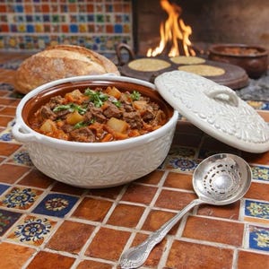 May include: A white ceramic serving bowl with a floral design, filled with a stew of meat and vegetables. A decorative lid sits beside the bowl, and a silver serving spoon rests on the tiled surface. A loaf of bread and a fire are in the background.