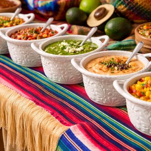 May include: A vibrant display of various salsas and dips in white, floral-patterned bowls, each with a spoon. The table is covered with a colorful striped textile with fringe. Limes, avocados, and tomatoes are also present, suggesting a Mexican cuisine theme.