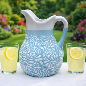 Puede incluir: Una jarra de cerámica con asa y boquilla blancas, y cuerpo azul claro con diseños florales blancos. Dos vasos de limonada con rodajas de limón frente a la jarra. La jarra mide aproximadamente 25 cm de alto.