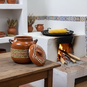 May include: A brown ceramic pot with a lid, decorated with green and white dots, sits on a wooden table. The lid is off, revealing the pot's interior. The pot has two black handles. A rustic kitchen scene includes a fireplace and other terracotta pottery.