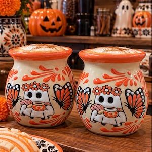 May include: Two round ceramic mugs with a cream-colored base and orange rims. Each mug features a hand-painted design of a white ghost wearing a flower crown and holding a small cup, flanked by orange and black monarch butterfly wings.
