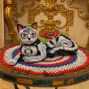 May include: A hand-painted ceramic cat figurine in a Day of the Dead style. The cat is black and white with colorful floral and skeletal designs. It rests on a round table with a crocheted doily.