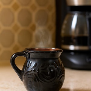 May include: A black ceramic mug with a floral design and a handle. The mug contains a hot drink, with steam rising. A coffee machine is visible in the background.