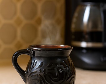 Può includere: Una tazza in ceramica nera con un motivo floreale decorativo e un manico. La tazza contiene una bevanda calda, con vapore che sale. Una macchina per il caffè è visibile sullo sfondo.