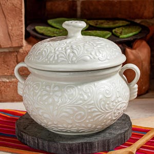 May include: A white ceramic casserole dish with a lid, featuring an embossed floral design. The dish has two handles and sits on a dark gray, round wooden base. The background includes a brick wall and a colorful striped cloth.