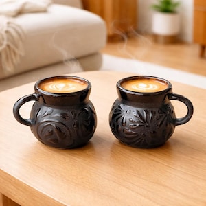 Może przedstawiać: Dwa czarne ceramiczne kubki z kwiatowymi wzorami, każdy wypełniony kawą i zwieńczony latte art. Kubki mają zaokrąglone korpusy, małe uchwyty i stoją na jasnym drewnianym stole. Para unosi się z kawy, sugerując, że jest gorąca.