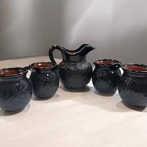 May include: A black ceramic jug and four matching mugs. The set has a dark, glossy finish with embossed floral patterns. The interiors of the mugs and jug are a contrasting brown colour. The set is displayed on a light-coloured surface.