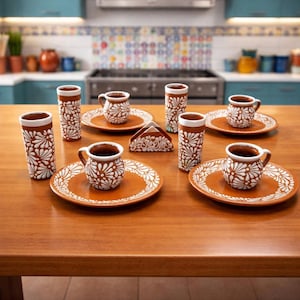 以下が含まれることがあります： テラコッタ色のセラミック食器セット。白い花の模様が特徴です。小さなマグカップ、お皿、背の高いグラスが含まれており、木製のテーブルに配置されています。お揃いのデザインのナプキンホルダーも付いています。