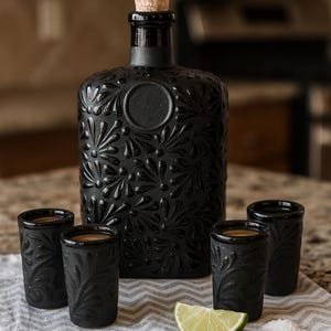 May include: A black, embossed liquor bottle with a cork stopper, alongside four matching shot glasses. A lime wedge and salt are on a patterned cloth, suggesting a tequila set.