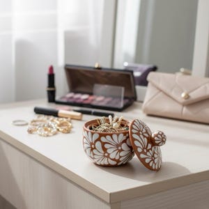 May include: A ceramic jewellery box with a lid, decorated with brown and white floral patterns. The box is open, displaying necklaces and bracelets. Other items on the table include makeup and a small purse.