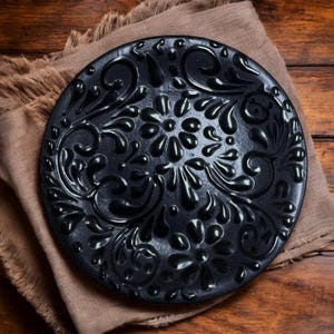 May include: A black decorative plate with an embossed floral design. The plate is round and sits on a brown cloth, with a wooden background. The plate's design includes flowers and swirling patterns.