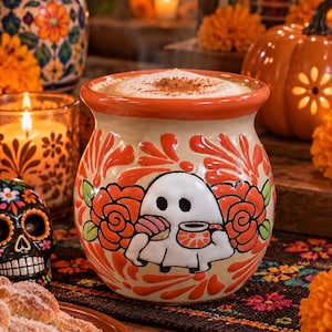 May include: A ceramic mug with a cream-colored base and a wide orange rim, featuring a hand-painted design of a white ghost holding a pastry and a cup, surrounded by orange floral patterns and green leaves, filled with a frothy beverage topped with cinnamon.