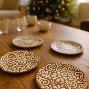 以下が含まれることがあります： 白い花柄と渦巻き模様が施された、茶色のセラミックコースター4枚セット。木製のテーブルに配置され、背景にはクリスマスツリーや食器がぼやけています。各コースターの直径は約10cmです。