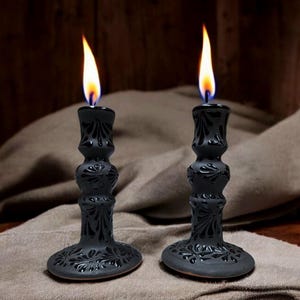 Pode incluir: Dois castiçais pretos com chamas acesas. Os castiçais apresentam um design floral detalhado e estão colocados sobre uma superfície neutra. As chamas são amarelas e laranja, contrastando com os castiçais escuros. Um acessório decorativo para casa.