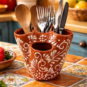 Op de afbeelding: Een terracotta keukengerei houder met witte bloemmotieven, met houten lepels, vorken en messen. Het staat op een kleurrijk betegeld oppervlak, geschikt voor een keuken. Het ontwerp is sierlijk en decoratief.