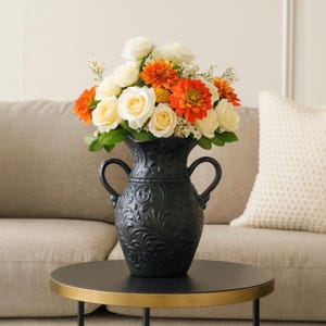 May include: A matte black ceramic vase with two side handles and an embossed floral scroll pattern, filled with a bouquet of white roses, orange dahlias, and small white filler flowers, creating a high-contrast floral arrangement.