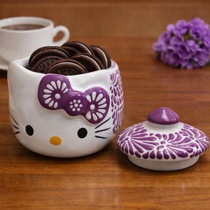 Peut inclure: Un pot à biscuits en céramique blanche en forme de visage de chat, avec un nœud papillon et des motifs floraux violets. Le pot est rempli de biscuits sandwich au chocolat. Le couvercle est à côté du pot. Une tasse de café et des fleurs violettes sont en arrière-plan.