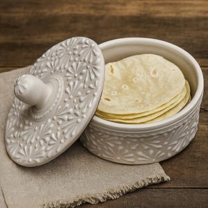 Puede incluir: Un calienta tortillas de cerámica blanca con tapa, con un diseño floral en relieve. La tapa está ligeramente entreabierta, revelando una pila de tortillas en su interior. Sobre una servilleta de lino beige, sobre madera.