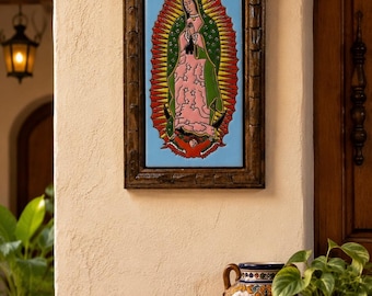 May include: A rectangular wall plaque featuring a depiction of Our Lady of Guadalupe in a dark wood frame. The image shows a figure in a pink robe and green mantle surrounded by a yellow and red sunburst on a light blue background.