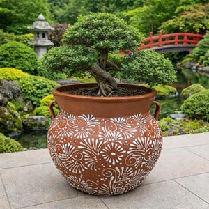 以下が含まれることがあります： 白い花の模様が入った、大きな茶色の陶器鉢に入った盆栽。 鉢には2つの取っ手があり、石の表面に置かれています。 背景には、赤い橋と緑豊かな緑がある日本庭園があります。