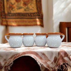 May include: Four white ceramic mugs with brown rims and intricate white floral designs. The mugs are arranged in a row on a white tablecloth with brown fringe.