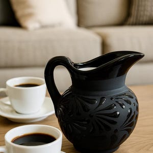 Może przedstawiać: Czarny ceramiczny dzbanek z kwiatowym wzorem, zakrzywionym uchwytem i dzióbkiem. Dwie białe filiżanki do kawy ze spodkami stoją na drewnianym stole. Kawa jest ciemna. W tle beżowa sofa.