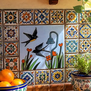 May include: A wall installation of square ceramic tiles featuring a central mural of two black swallows flying over orange flowers and a blue bell-shaped flower. The surrounding tiles display intricate blue, orange, and yellow floral patterns.