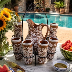 May include: A terracotta pitcher and five matching tall tumblers featuring intricate white floral and swirl patterns, held in a black metal wire caddy. The set is crafted from reddish-brown clay with a smooth finish and white painted details.