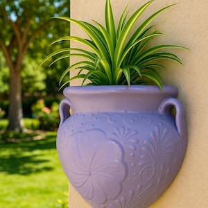 Puede incluir: Una maceta de pared de color lavanda con un diseño floral y dos asas, que contiene una planta de rayas verdes y blancas. La maceta está montada en una pared beige, con un fondo verde borroso.