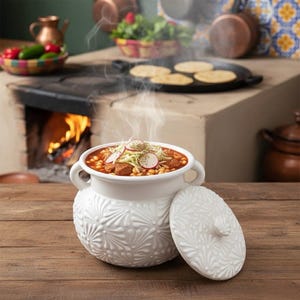Può includere: Una pentola di servizio in ceramica bianca con coperchio, piena di uno stufato fumante, guarnito con verdure. La pentola ha un design floreale in rilievo. Lo sfondo include un forno a legna, tortillas e pentole di rame.