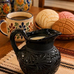May include: A black ceramic pitcher with a glossy finish and a matte base, featuring a raised floral pattern on the body. The pitcher has a curved handle and a pouring spout, and it is filled with a light-colored liquid, likely milk or cream.
