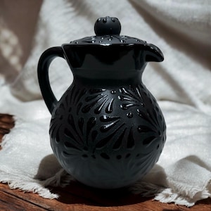 Op de afbeelding: Zwarte keramische kan met een deksel en een ingewikkeld bloemmotief. De kan heeft een rond lichaam en een tuit aan de zijkant.